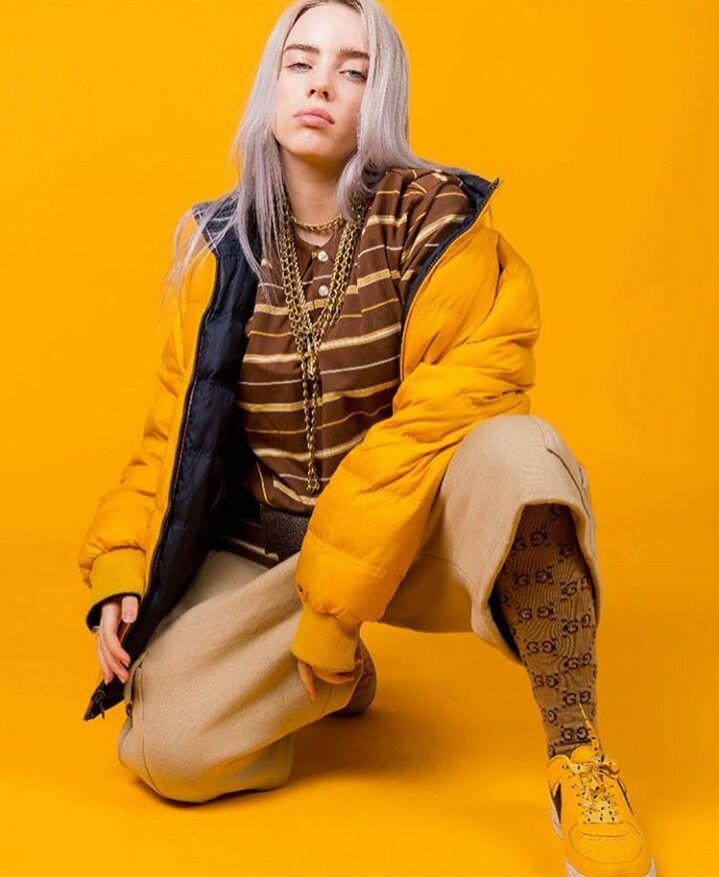 Billie Ellish is wearing a yellow jacket. She has on a brown and yellow striped shirt. She has on tan pants and brown socks with yellow shoes. The background is yellow