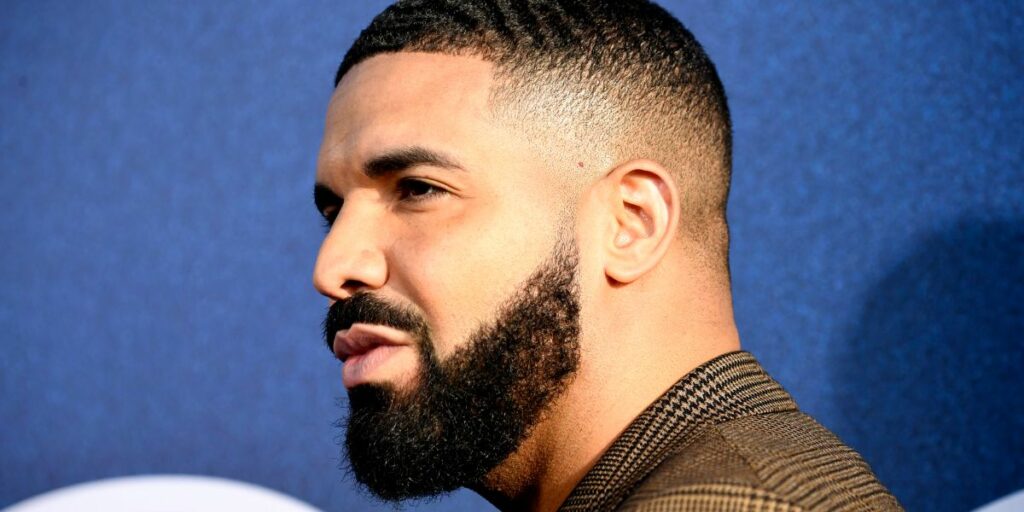 Drake is standing in front of blue background. Drake has on a plaid jacket.